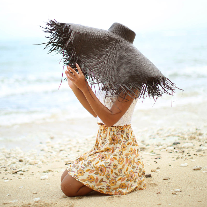 A woman is wearing BrunnaCo’s Amora Handwoven Frayed Oversized Straw Hat in black made by artisans in Bali. This tropical island hat is made for a perfect holiday vacation and coastal getaways. The photograph is taken by Faruq Adib’s team, Matarasa in Bingin Beach, Bali.