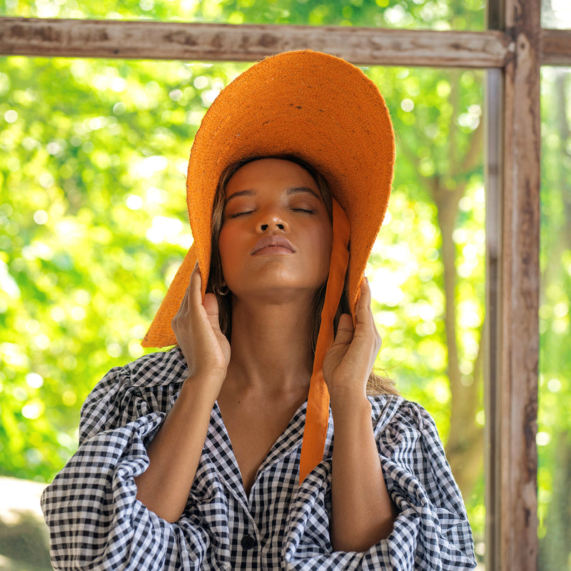 LOLA Wide Brim Jute Straw Hat In Orange. Braided meticulously by artisans in Gianyar, Bali from natural Jute material, this sweet hat offers both comfort and elegance. Designed with ideal thickness and weight that allow its brim to be easily folded, stretched, and styled, making it easy to take to any holiday trips or summer vacation.