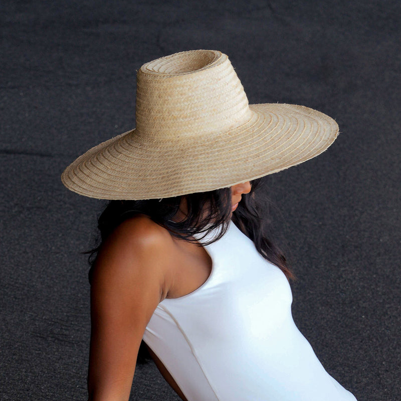 A woman is wearing BrunnaCo Rianna Straw hat made from locally harvested palm straw in Bali. This versatila hat is perfect for a day at the beach or a stroll around town on a sunny summer day