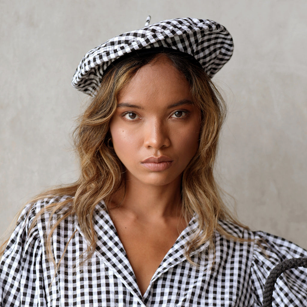 A woman is wearing BrunnaCo Simone Beret hat and she's ready for a casual stroll at the beach. The parisian beret hat has gingham pattern in black and white color